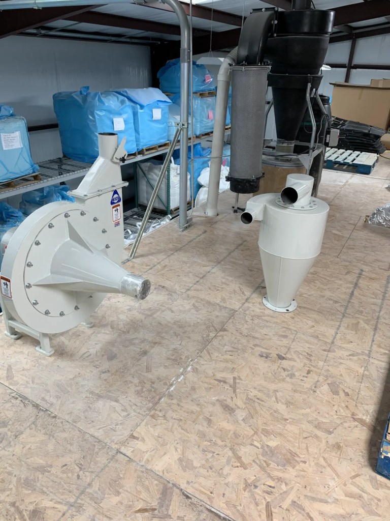 Industrial equipment in a storage area, featuring a large white machine with a conical end and a cylindrical component, surrounded by blue bags of material.